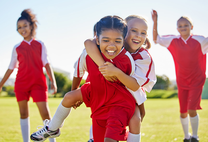 Happy young girls in red soccer uniforms playing and giving piggyback rides on a sunny field, highlighting how exercise and teamwork can benefit children with ADHD.