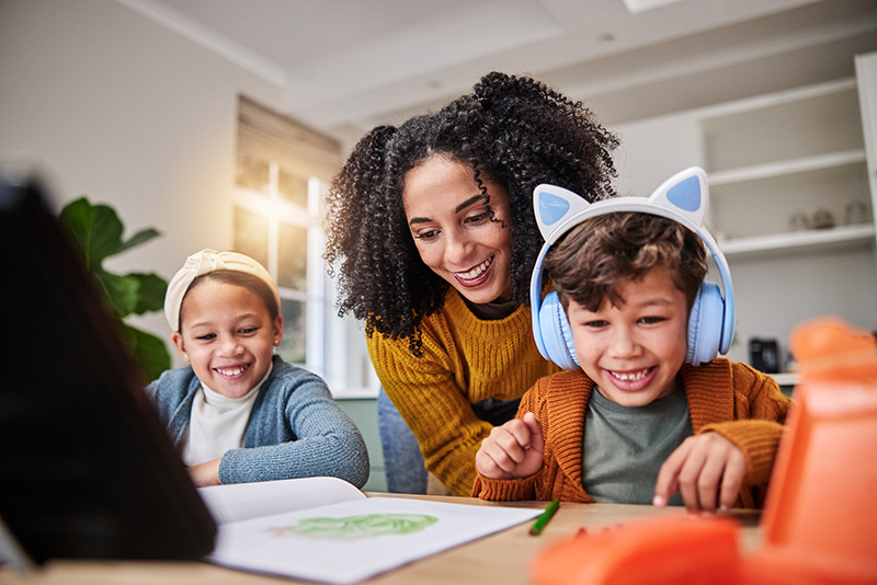 Smiling mother helping two children with homework and creative activities at home, with one child wearing headphones, promoting learning and focus for kids with ADHD.