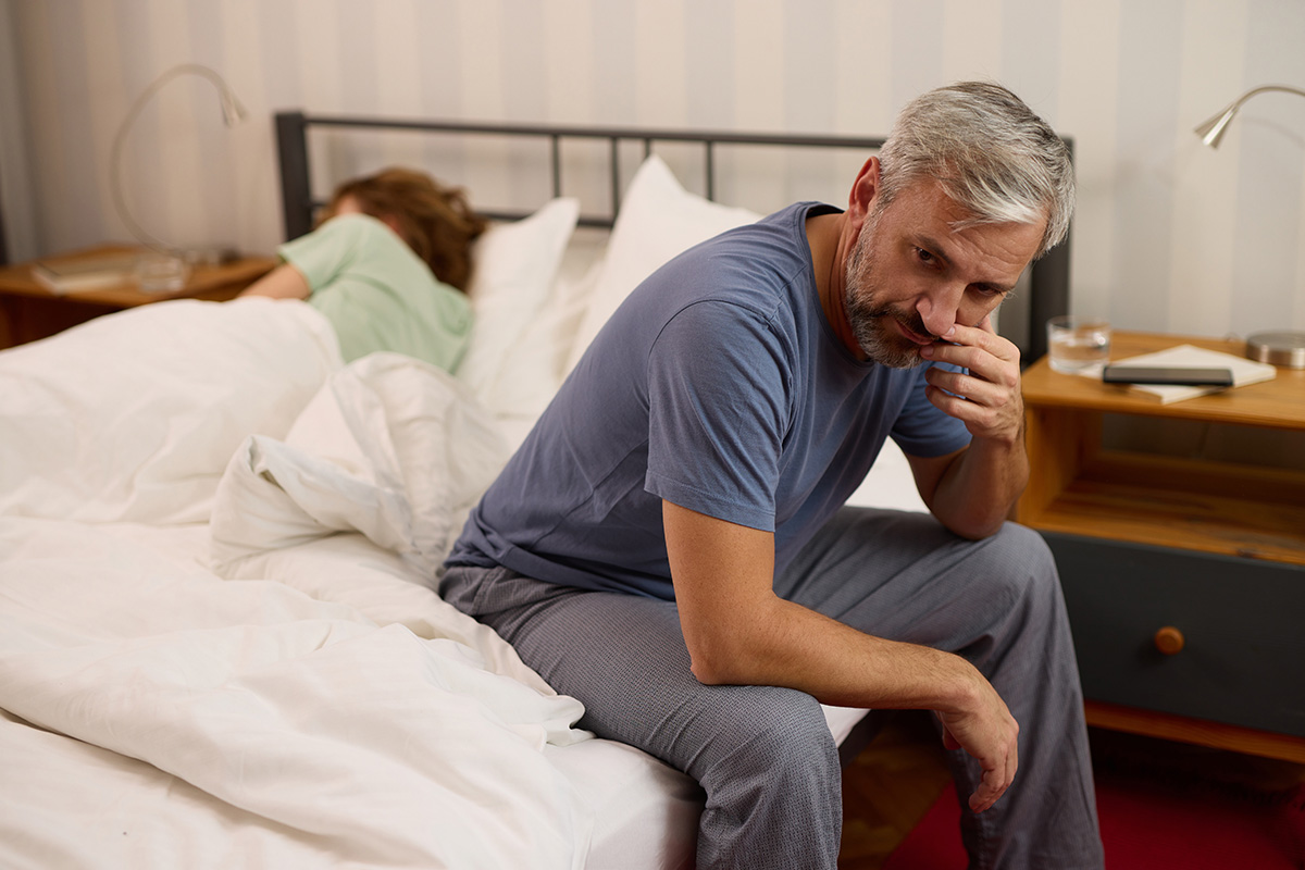 Middle-aged man sitting on the edge of a bed, appearing thoughtful and concerned, with a sleeping partner in the background, symbolizing relationship or personal challenges.