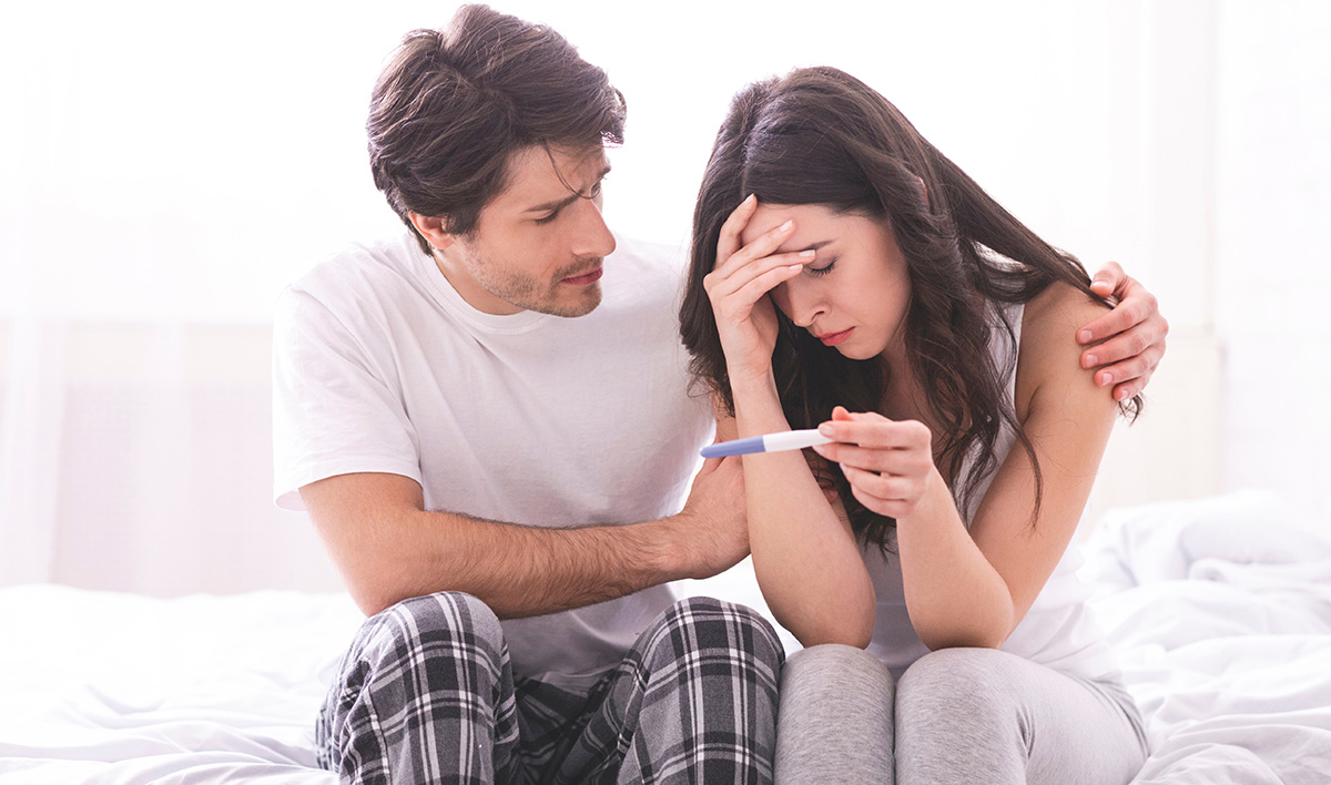 Couple sitting together on a bed, the man comforting a distressed woman holding a negative pregnancy test, symbolizing emotional support and challenges related to infertility or family planning.